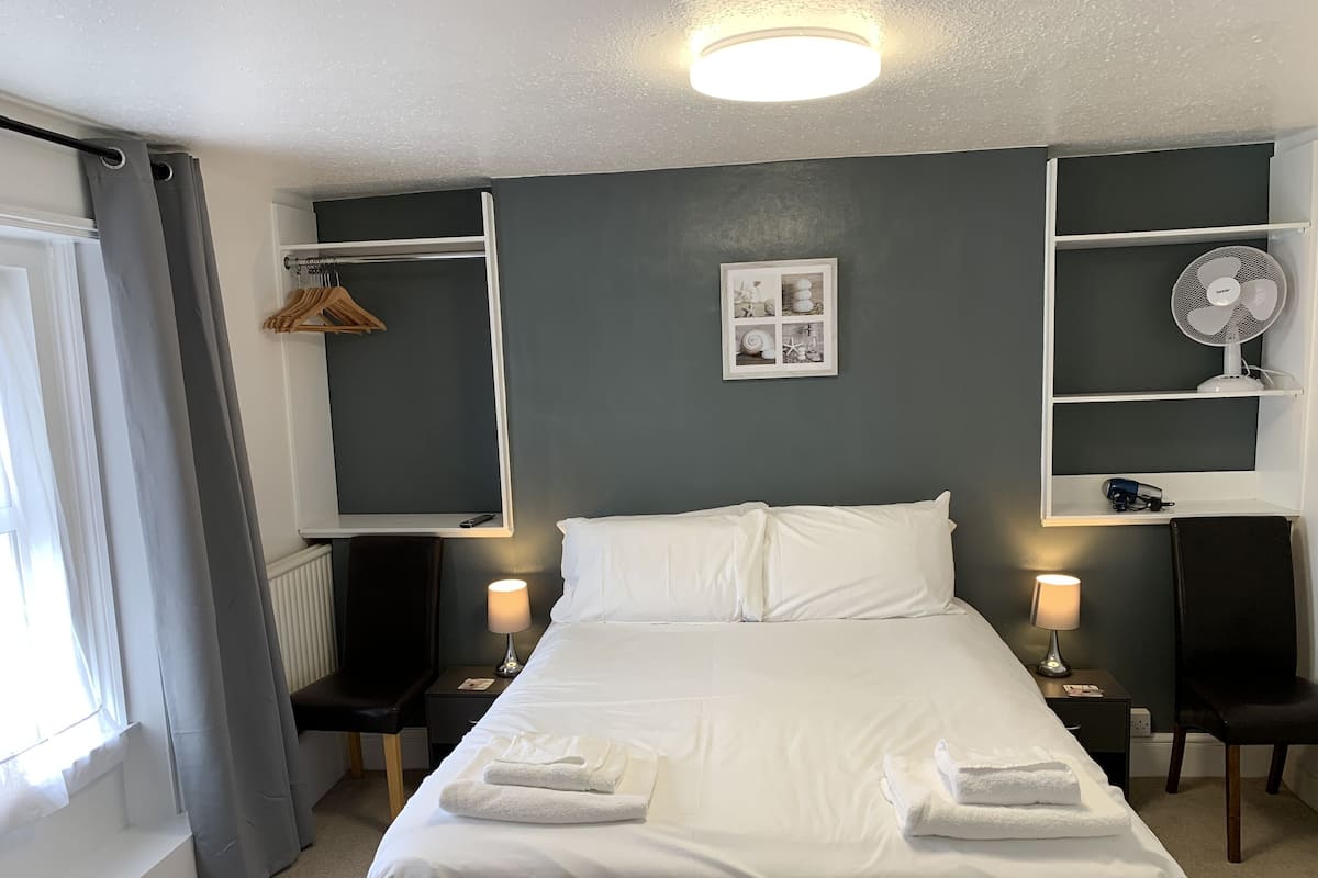 A neatly made bed with white linens and towels, a bedside table with a lamp, a wall-mounted shelf with a fan, and a framed picture on the wall.