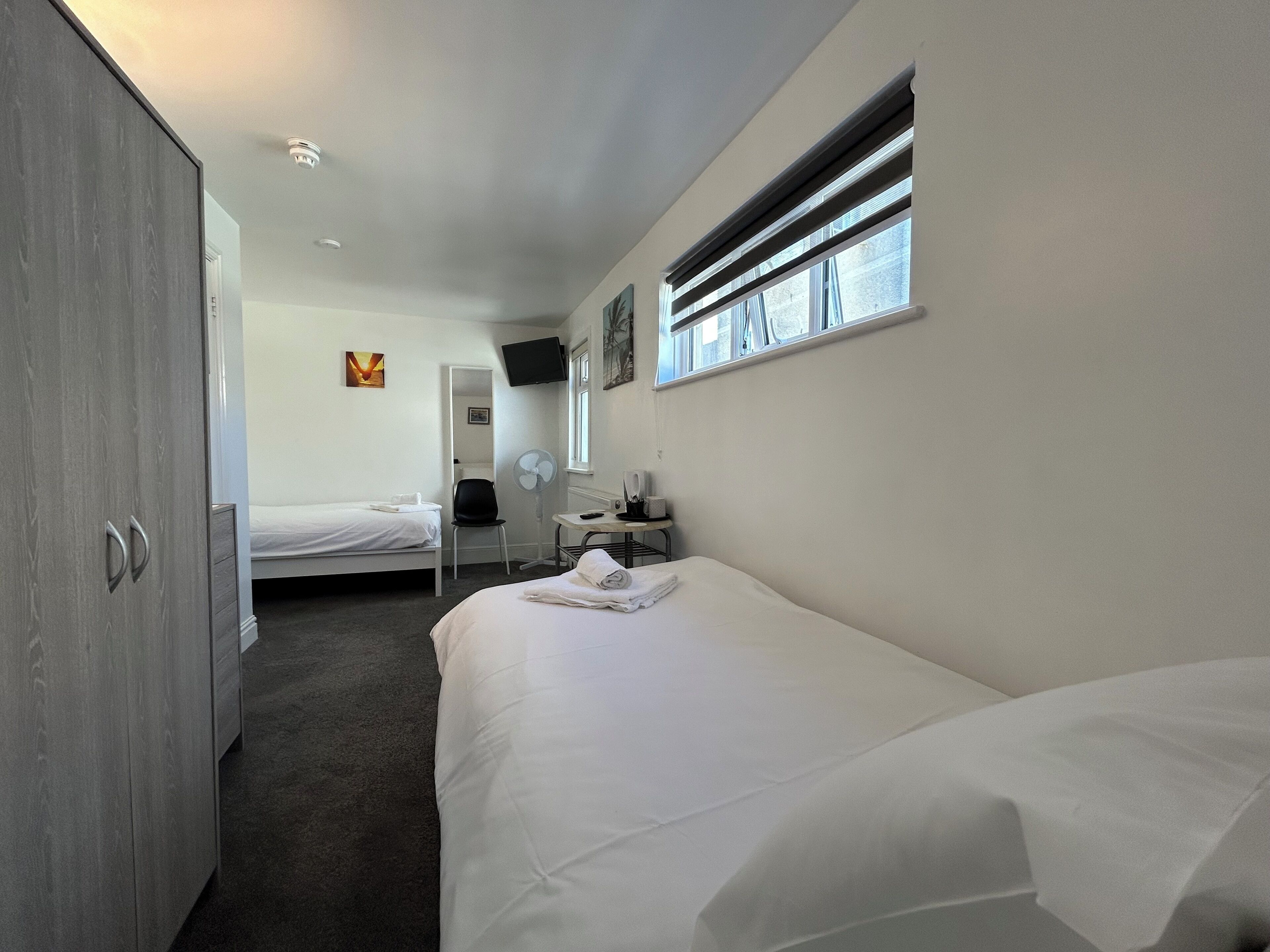 A hotel room with two beds, a desk with a chair, a wardrobe, a flat-screen TV, and a window with blinds.