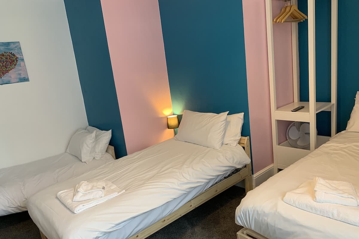 A room with two single beds, a wooden nightstand with a lamp, and a shelf with a fan.