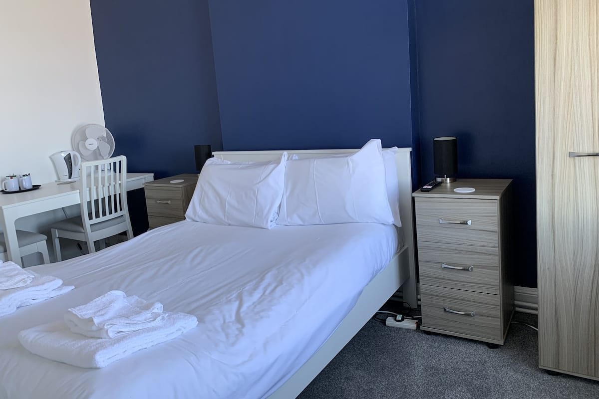 A neatly made bed with white linens and towels, a desk with a chair, a bedside table with a lamp, and a wardrobe.
