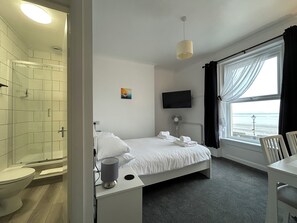 Una habitación de hotel con cama, televisor, ventana con cortinas, ducha y una mesita con lámpara.