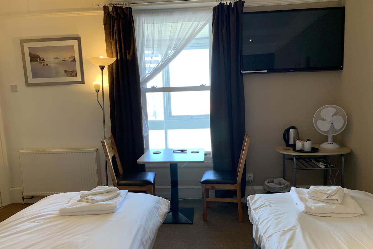 A hotel room with two beds, a table, chairs, a TV, a fan, and a window with curtains.