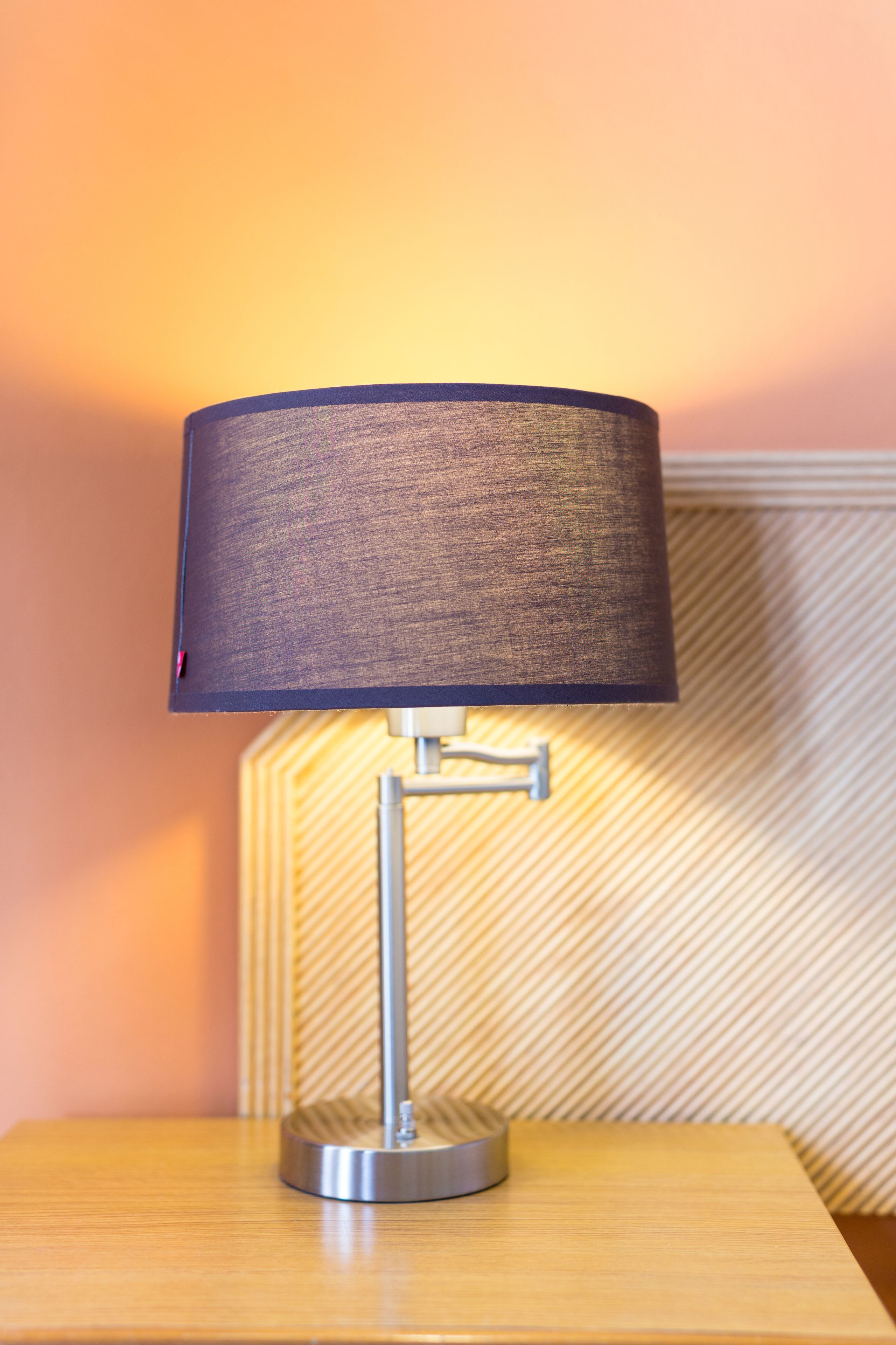 A modern table lamp with a cylindrical shade on a wooden table against a patterned wall.