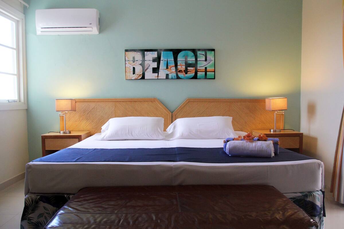 Hotelkamer met een bed, twee nachtkastjes met lampen, een wandairconditioner en een groot kunstwerk met de tekst 'BEACH'.