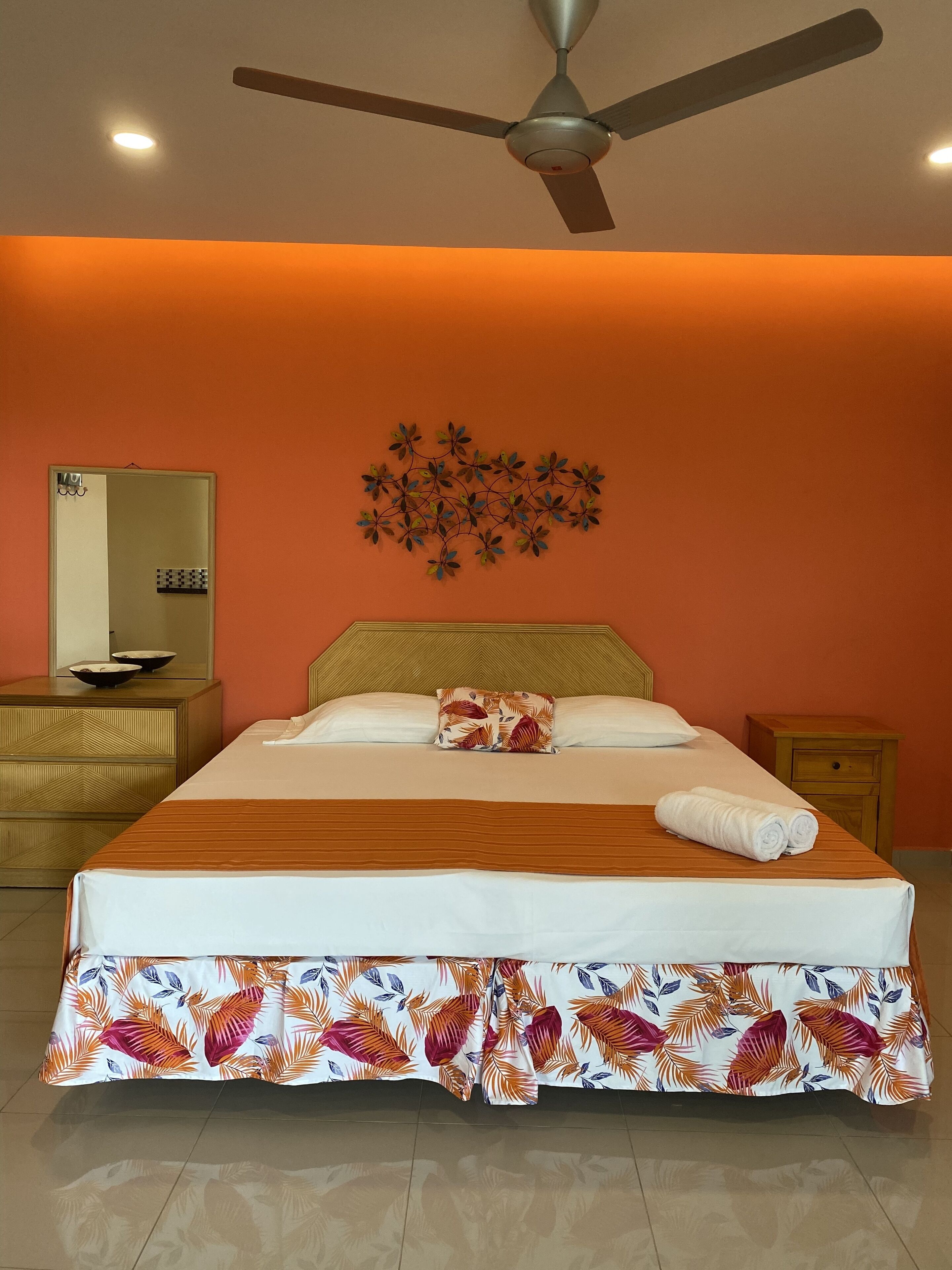 A hotel room with a bed, a ceiling fan, a wall-mounted mirror, and a decorative wall piece.