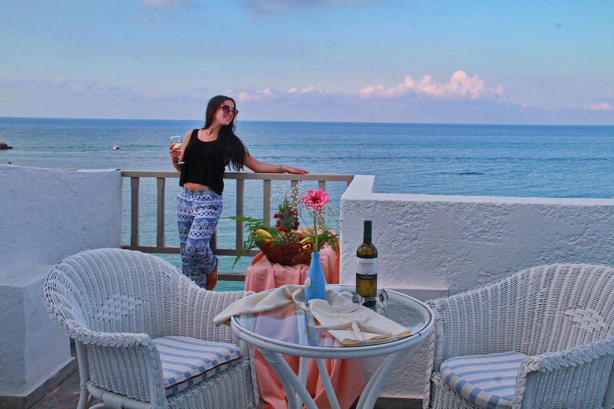 A balcony with wicker chairs, a table with a vase and a bottle, and a view of the sea.