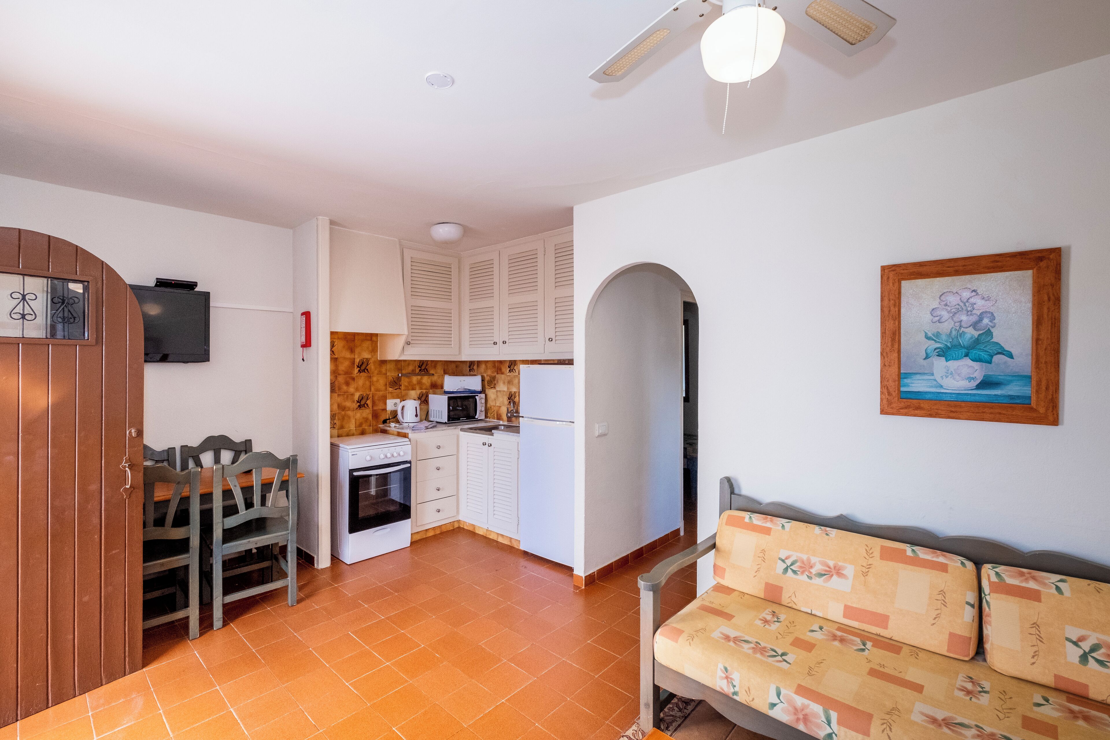 Cocina pequeña con armarios blancos, microondas y estufa. Cuenta con una mesa de comedor con sillas y un sofá con cojines estampados.