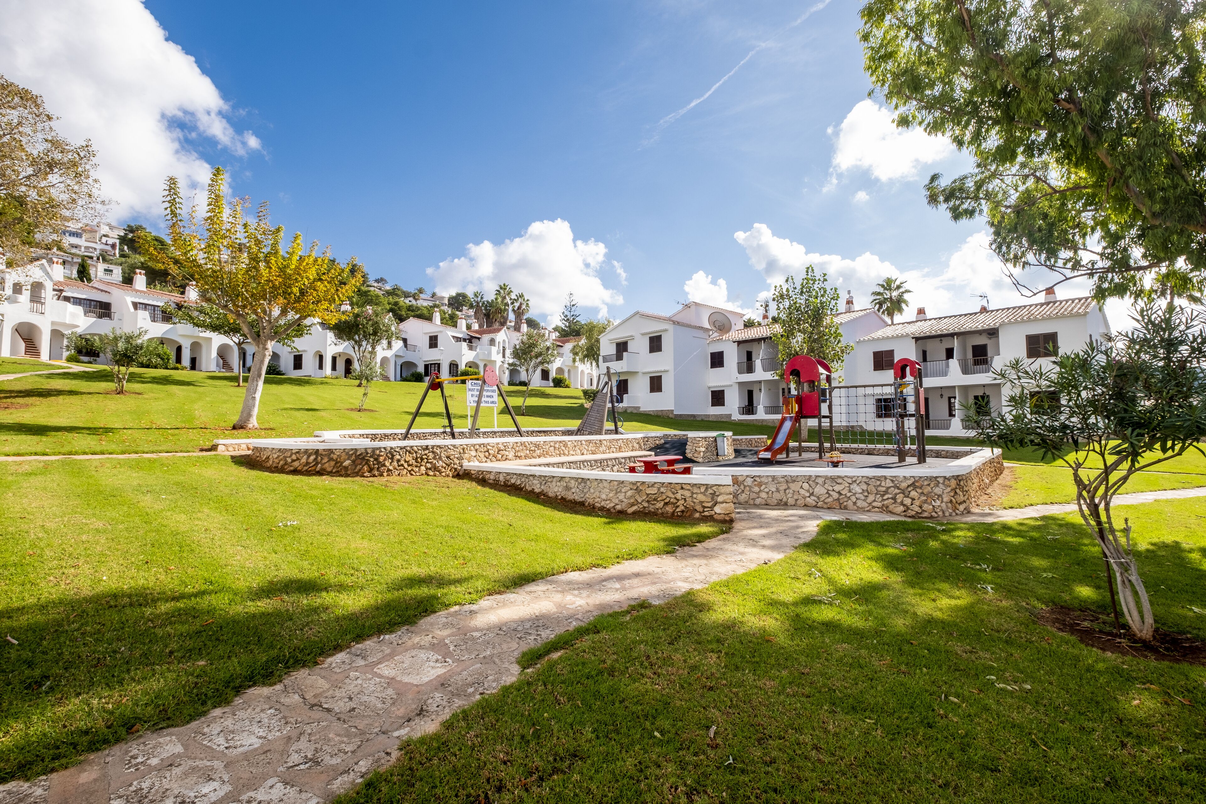 Un patio con un sendero de piedra, un parque infantil y edificios blancos con varios balcones.