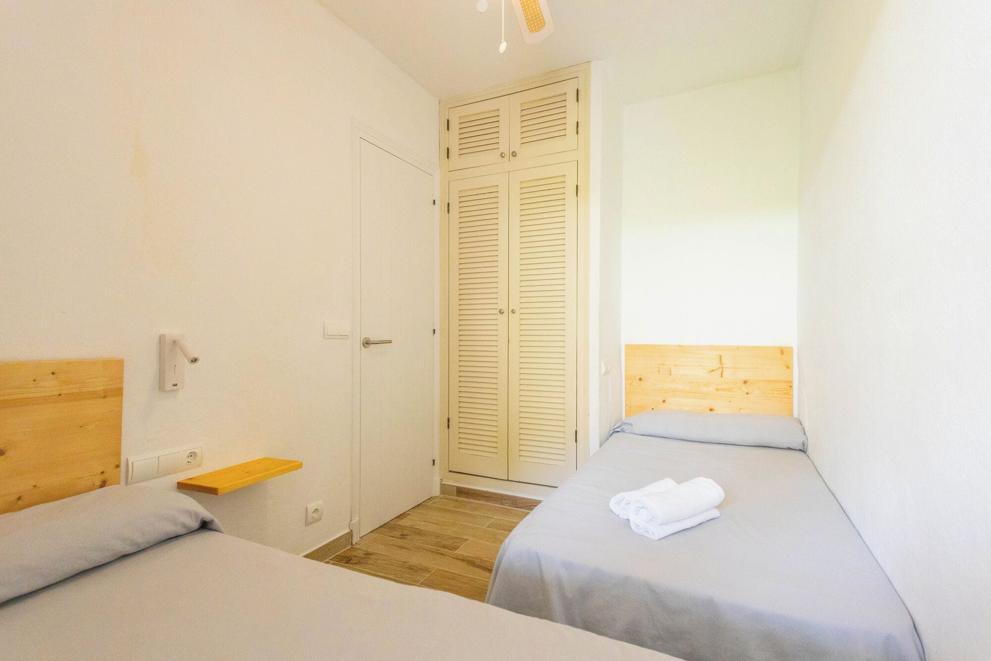 A small, clean room with two beds, a wooden headboard, a small shelf, and a door with louvered shutters.