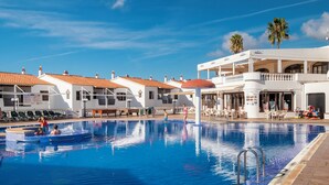 A swimming pool with a tiled deck, lounge chairs, and a building with a balcony. - Apartamentos Son Bou Gardens (Alayor)