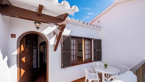 A white building with a covered patio area, a small table with chairs, and a door leading to an interior space. - Apartamentos Son Bou Gardens (Alayor)