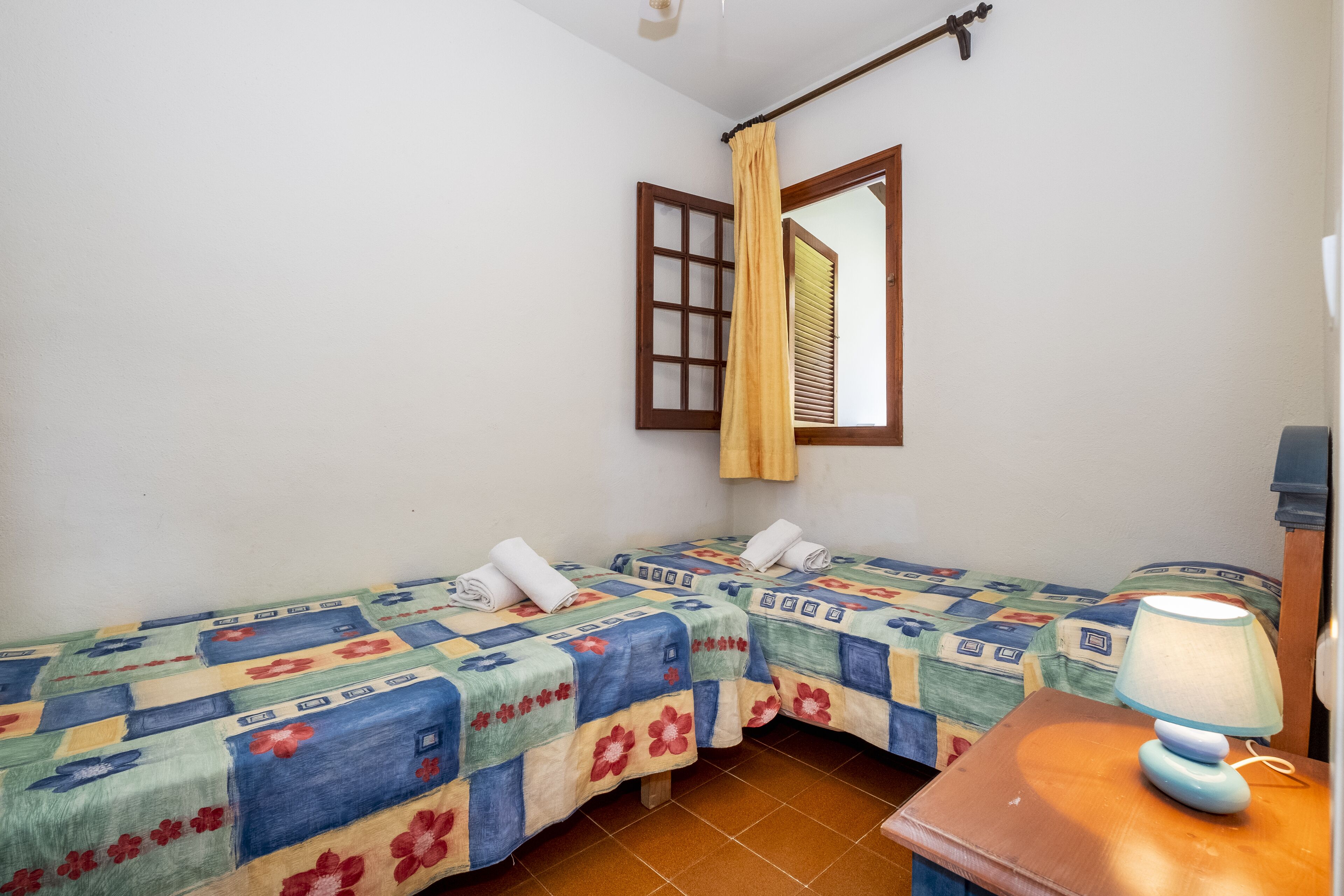 A room with two beds, each with a floral-patterned bedspread, a wooden nightstand with a lamp, and a window with a curtain.