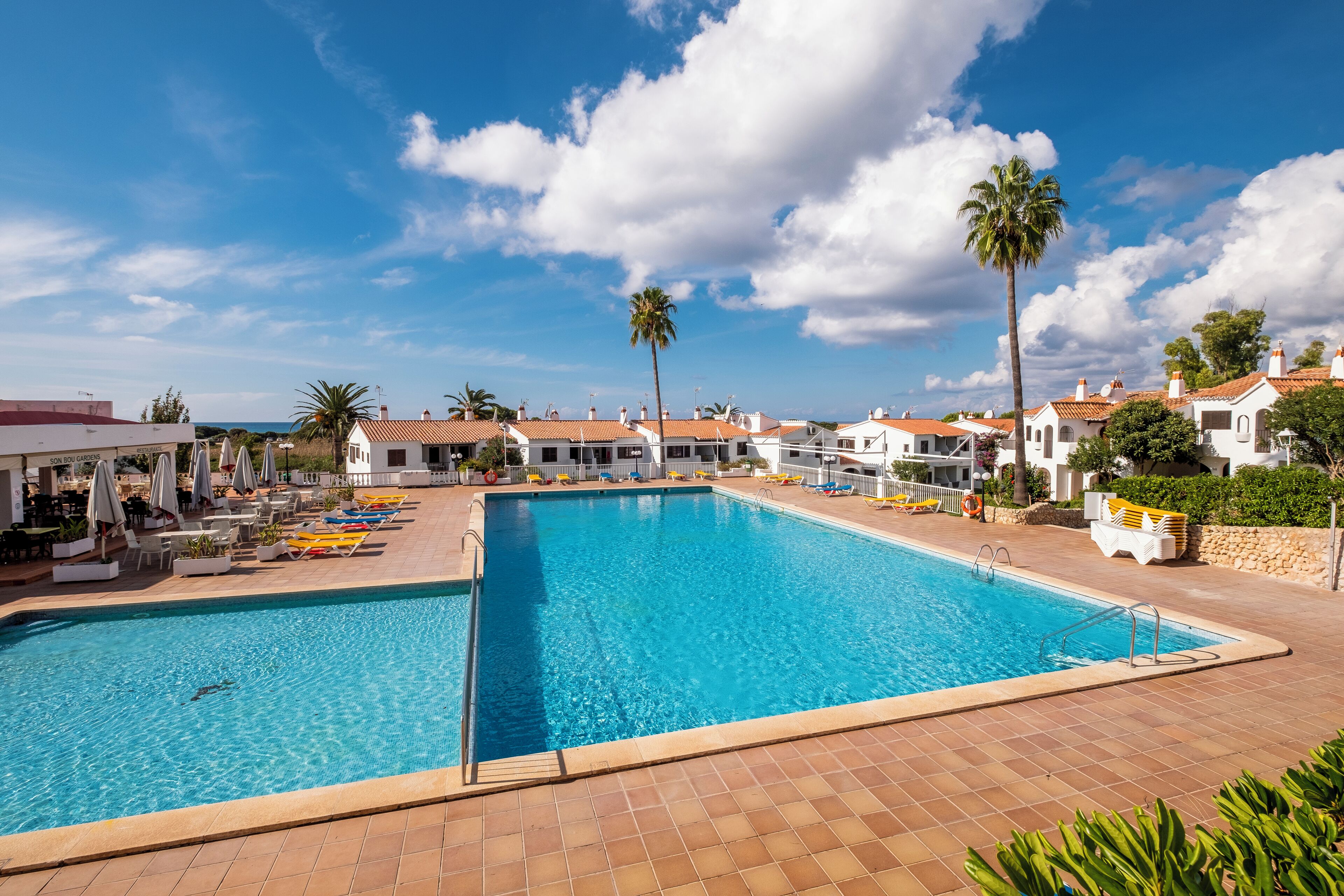 A swimming pool with lounge chairs and umbrellas, surrounded by white buildings with red roofs.