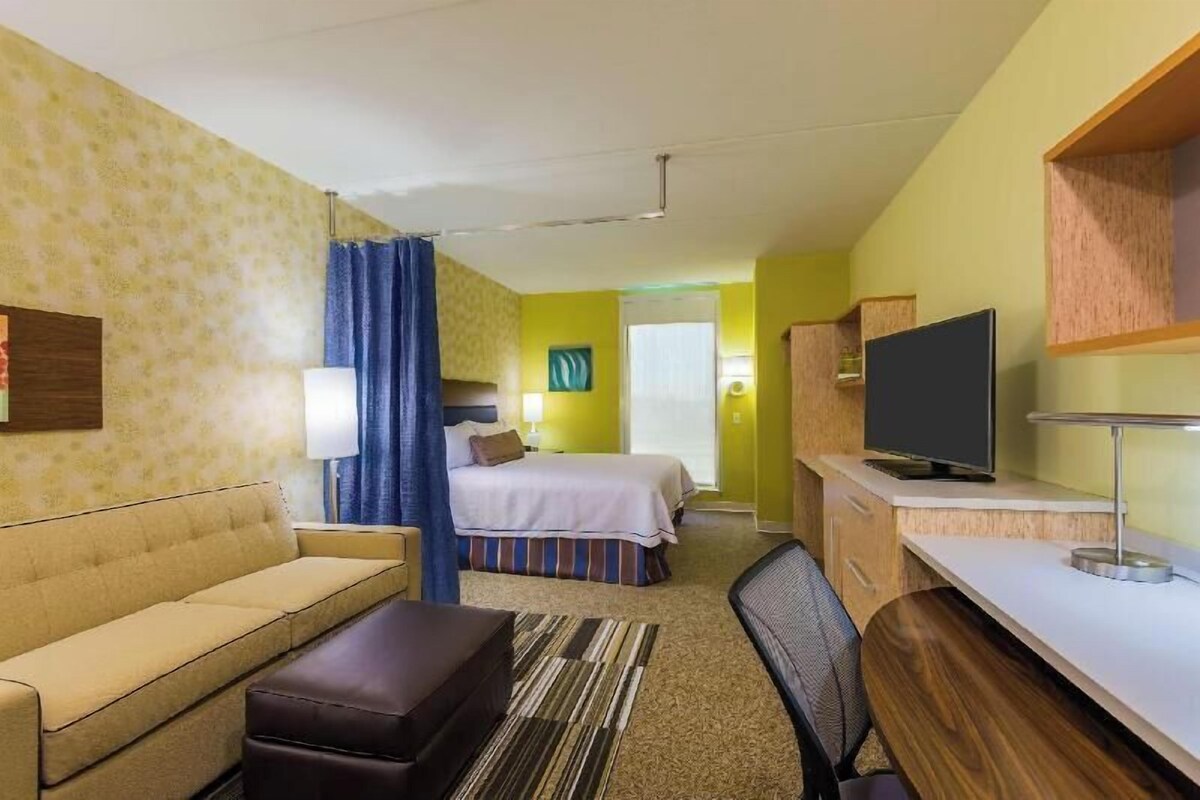 A hotel room with a bed, a sofa, a desk, a TV, and a bathroom visible in the background.