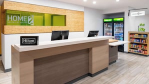 A hotel reception area with a reception desk, a 'Hilton Honors' sign, a 'HOME 2' sign, a 'really cold' and 'cold' sign above a refrigerator, and a shelf with snacks.