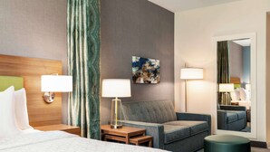 A hotel room with a bed, two bedside tables, a sofa, a round ottoman, and a mirror. - Home2 Suites by Hilton Lubbock (Lubbock)