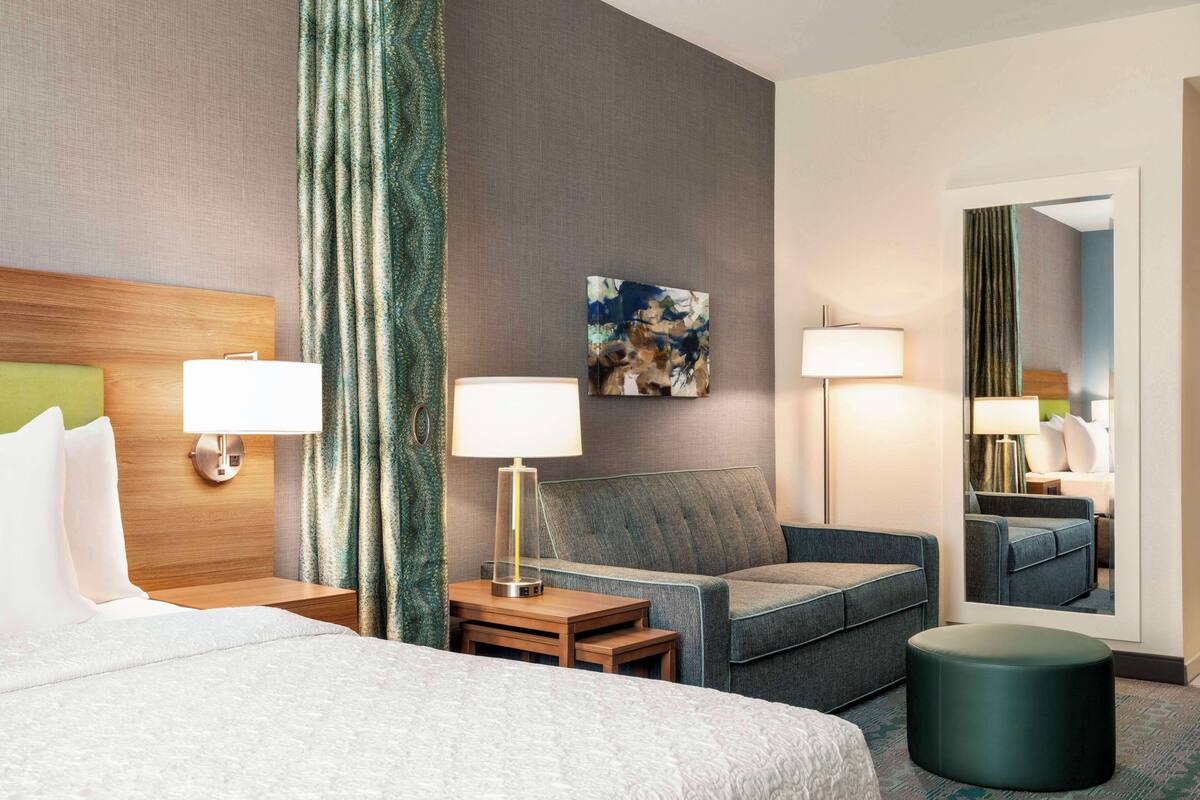 A hotel room with a bed, two bedside tables, a sofa, a round ottoman, and a mirror.