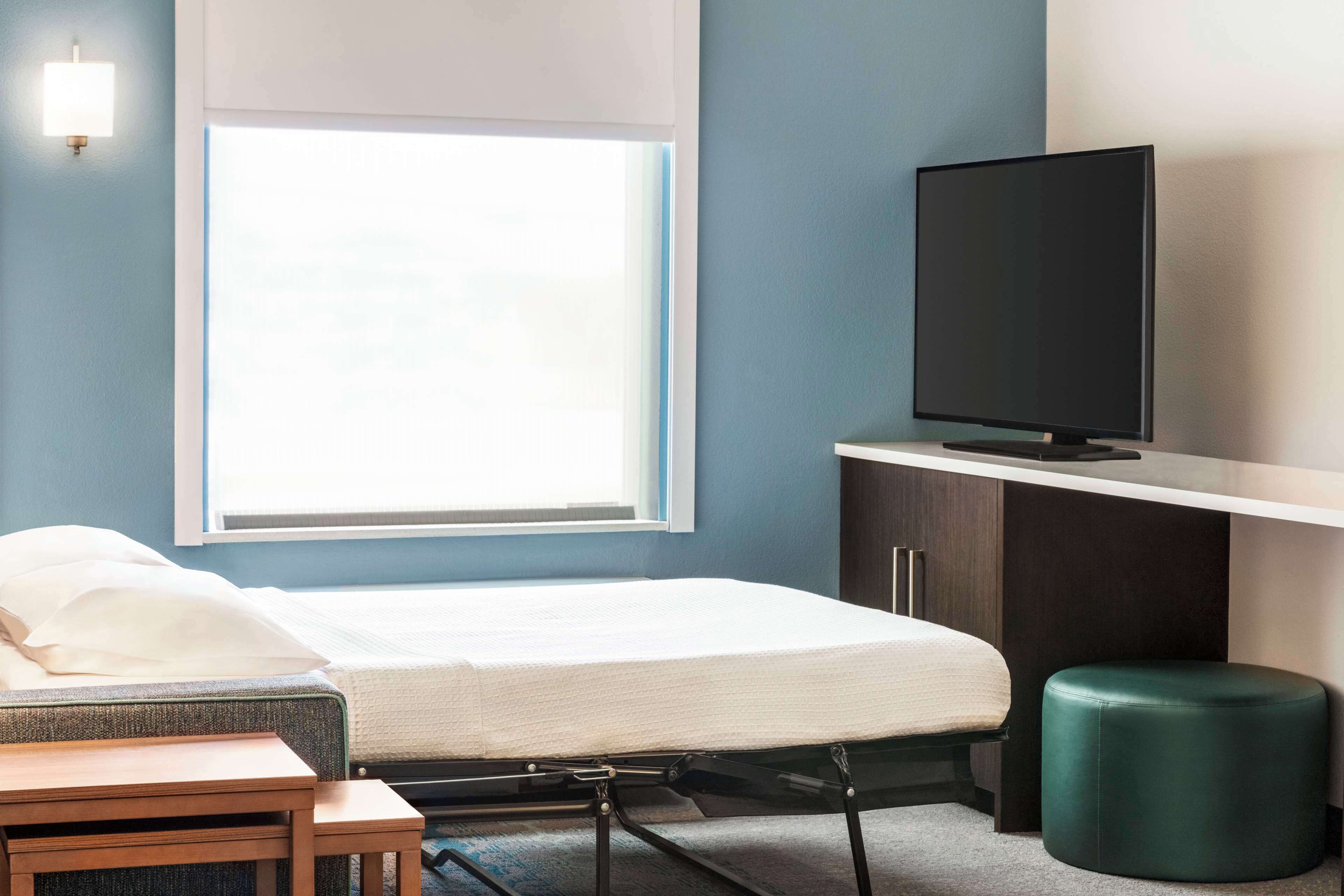 Una habitación de hotel pequeña con una cama, una mesita, un otoman verde, un televisor sobre un mueble bajo y una ventana que proporciona luz natural.