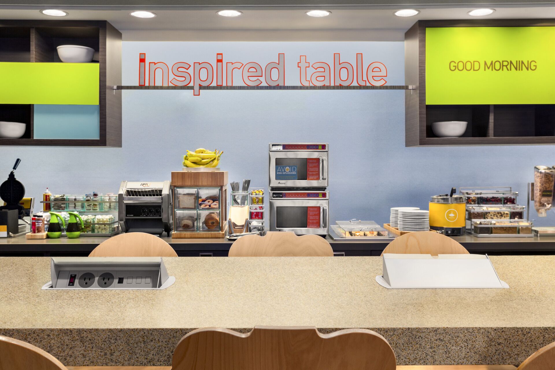 A breakfast area with a sign reading "Inspired table" and "GOOD MORNING". There are food dispensers, a fruit display, and seating with a notepad on the table.