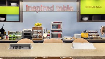 A breakfast area with a sign reading "Inspired table" and "GOOD MORNING". There are food dispensers, a fruit display, and seating with a notepad on the table.