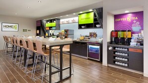A modern hotel lobby with a long communal table, bar stools, a coffee station, and a trash bin. - Home2 Suites by Hilton Lubbock (Lubbock)