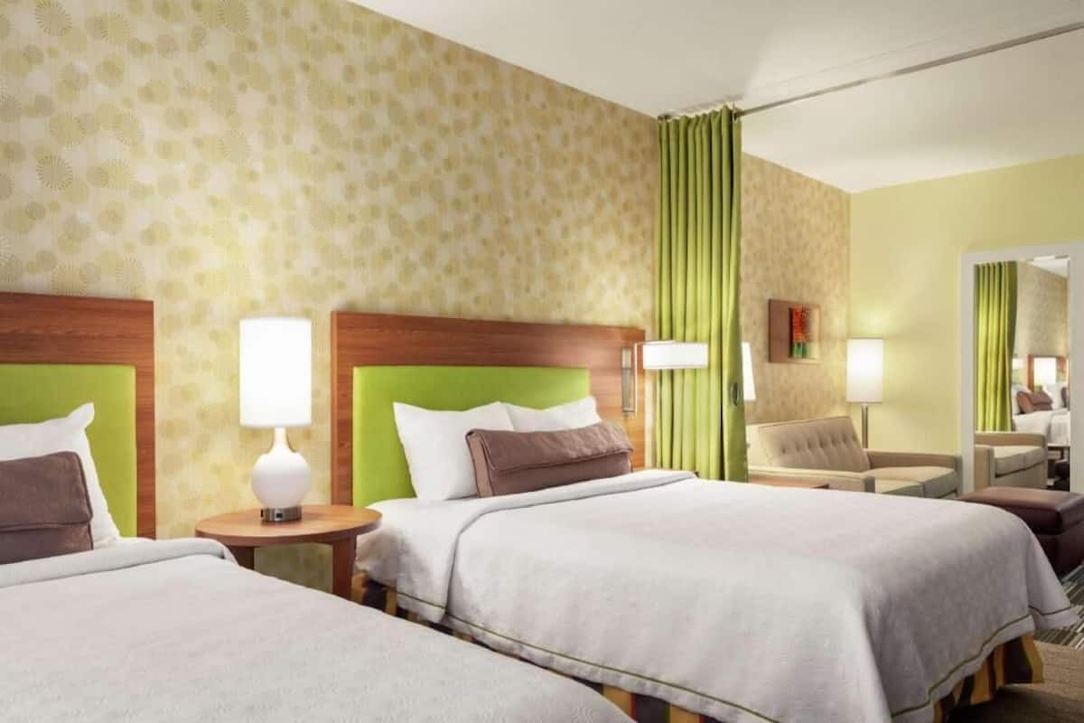 A hotel room with two beds, a green headboard, a mirror, a chair, and a lamp.