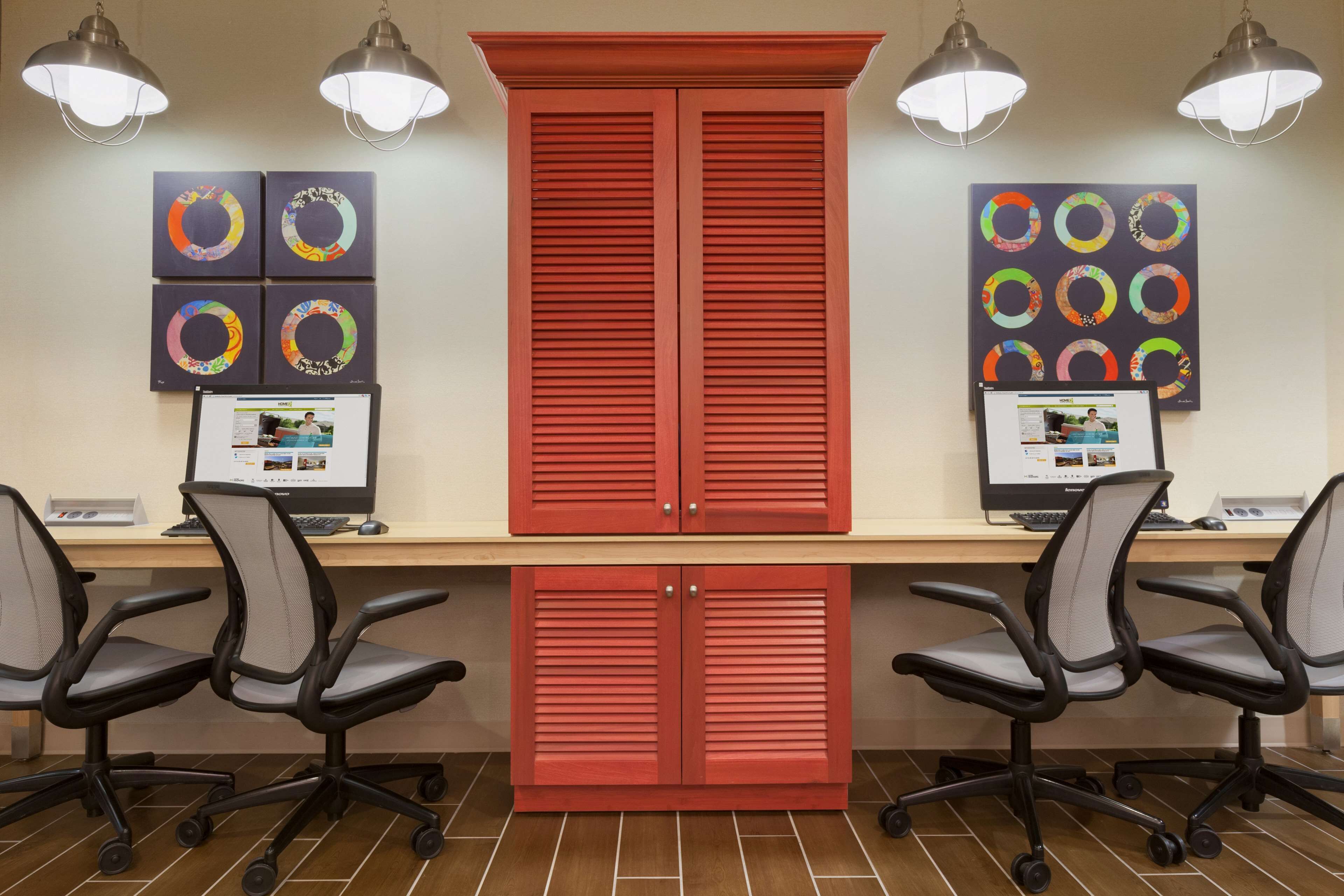 Un espacio de trabajo con dos escritorios, cada uno con una computadora y una silla. Hay dos armarios rojos y una pared decorada con obras de arte circulares.