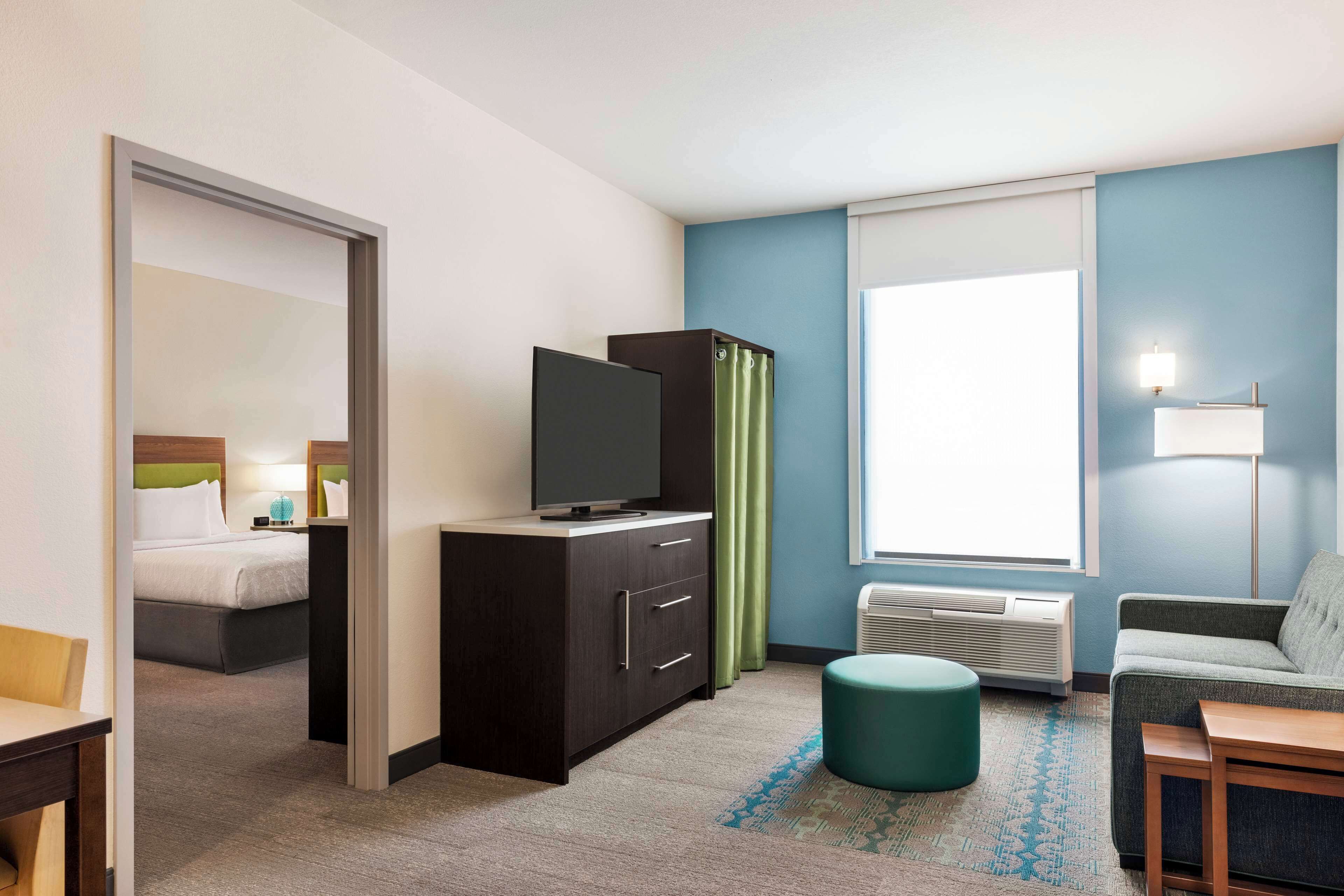 A hotel room with a bed, a TV on a cabinet, a sofa, a small table, and a window with curtains.
