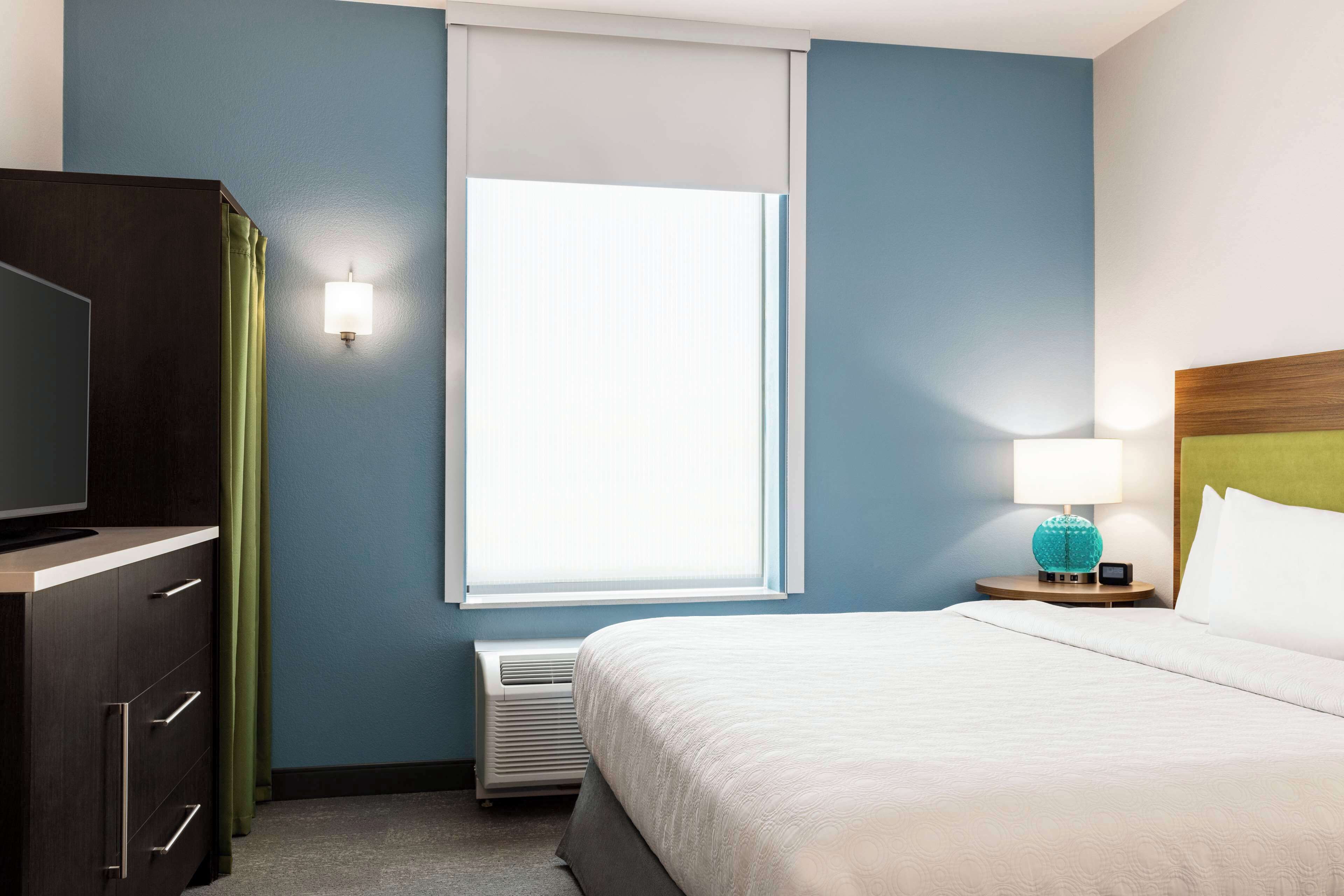 Een hotelkamer met een bed, een dressoir, een nachtkastje met lamp en een raam met jaloezieën.