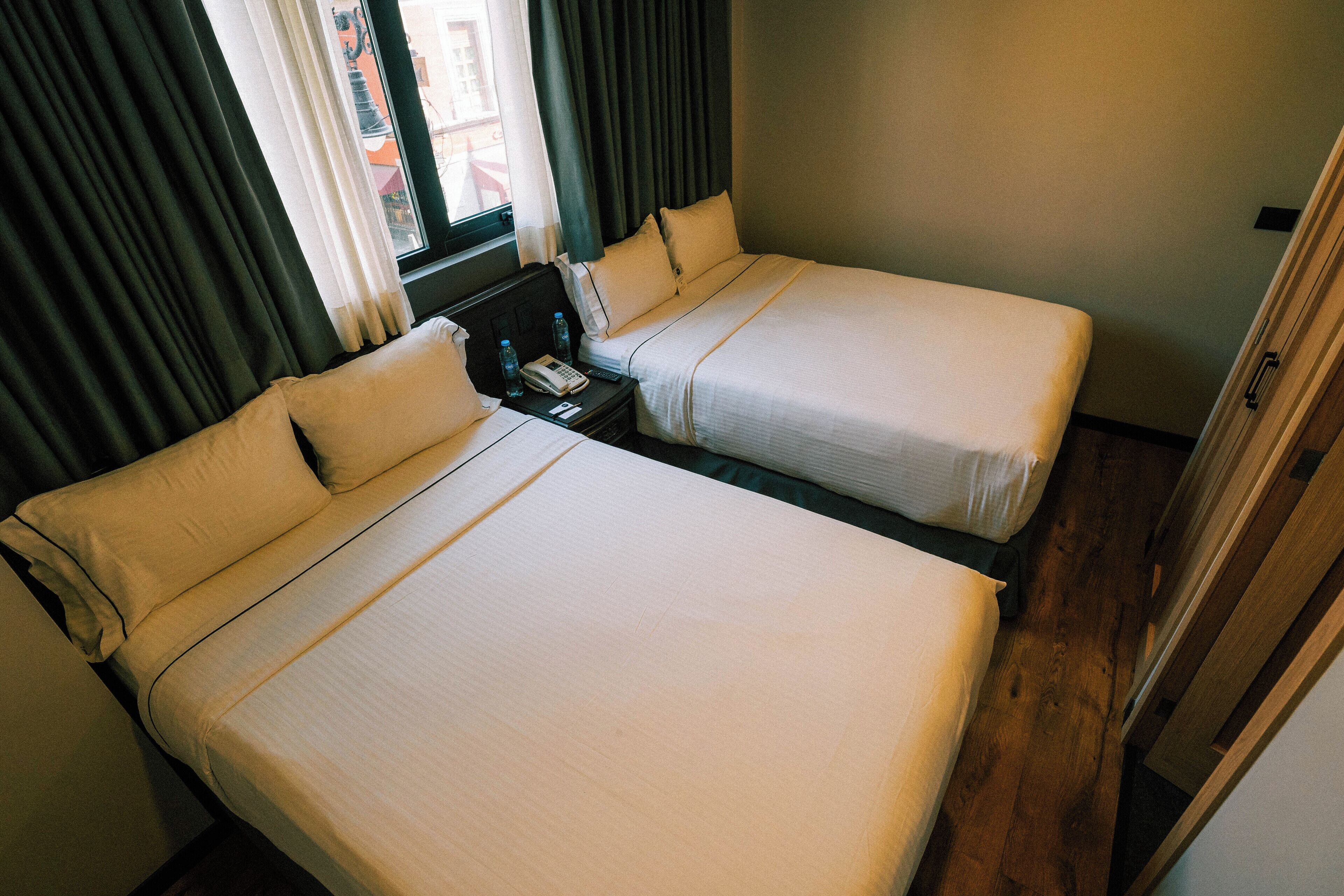 Habitación de hotel con dos camas, una mesita de noche, un teléfono y una ventana con cortinas.