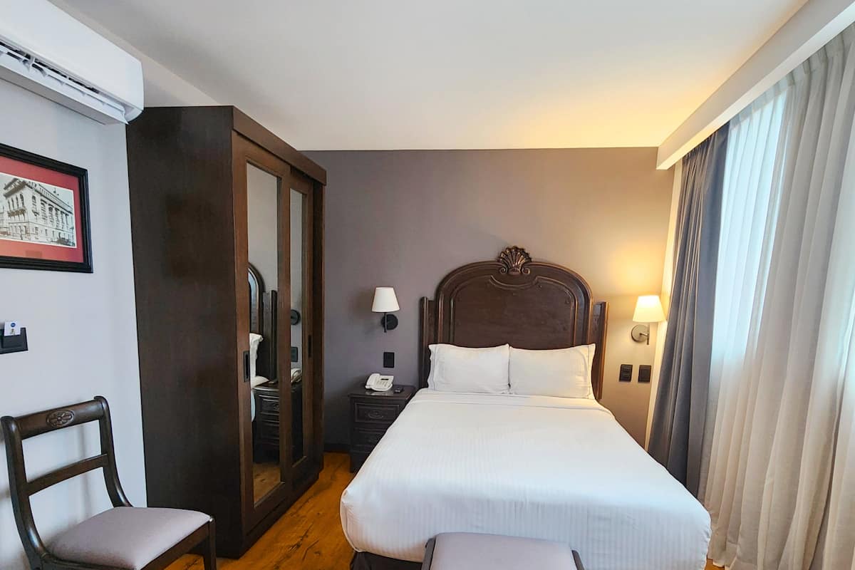 A hotel room with a large bed, two chairs, a wardrobe, a bedside table with a lamp, and a framed picture on the wall.
