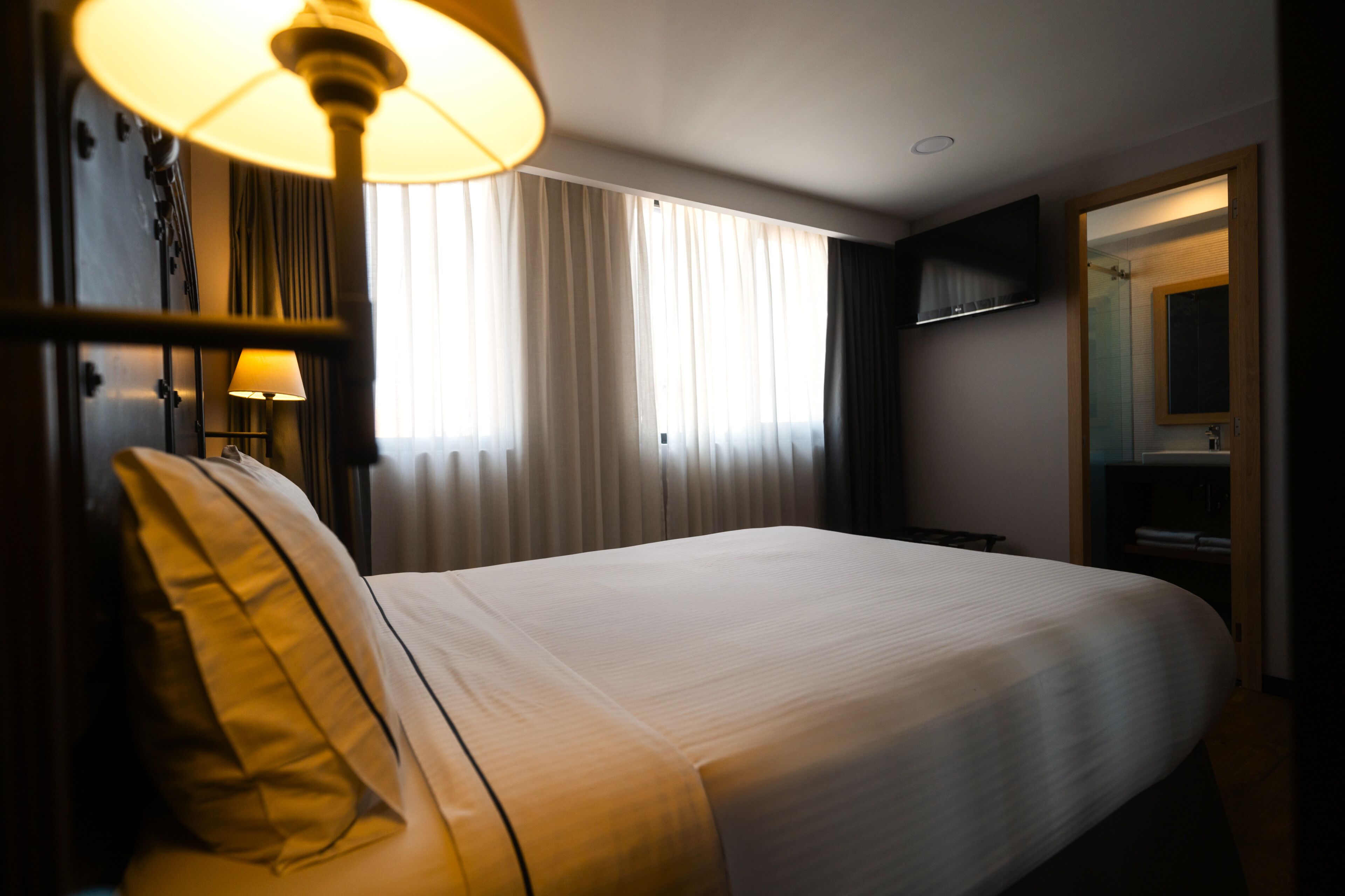 Een hotelkamer met een groot bed, een bedlampje, een aan de muur bevestigde televisie en een raam met gordijnen.