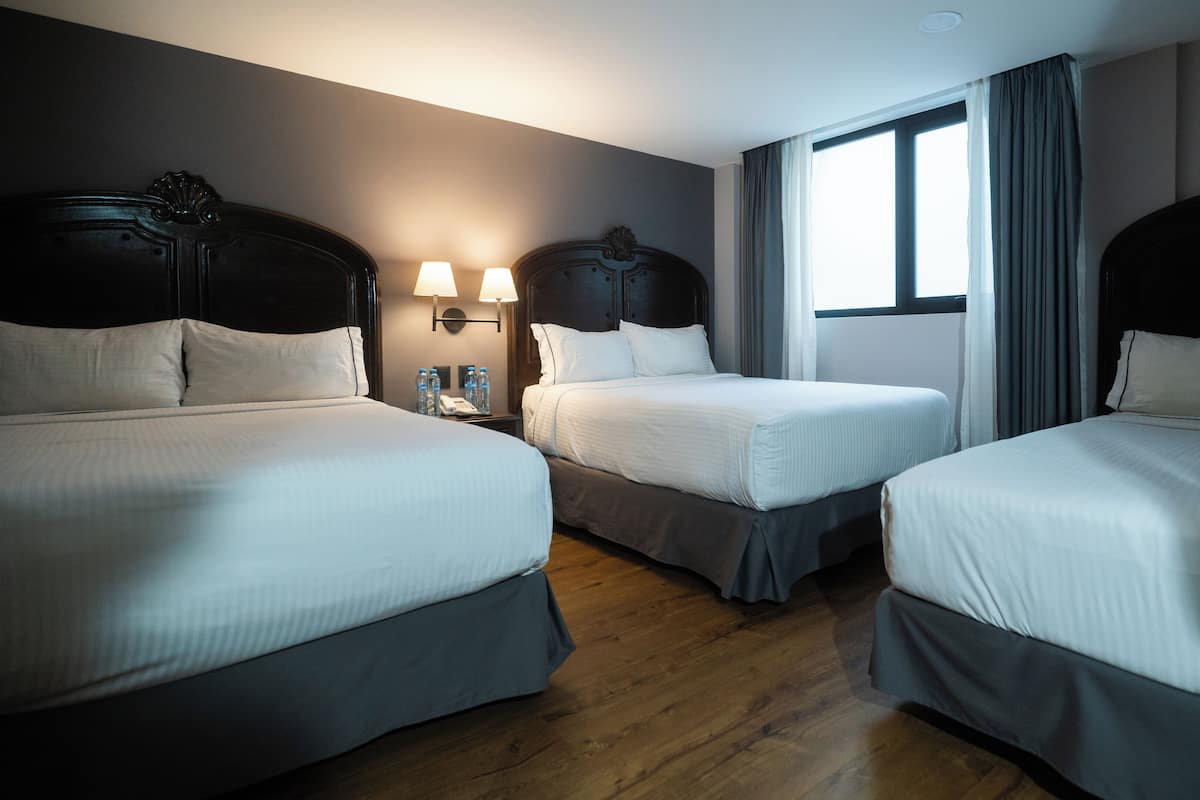 A hotel room with three single beds, a nightstand with a lamp, and a window with curtains.