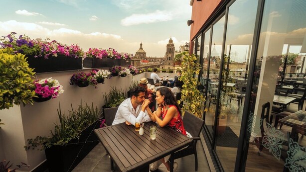 Una pareja cenando en una mesa al aire libre con vistas a la ciudad, rodeada de macetas con plantas y flores.