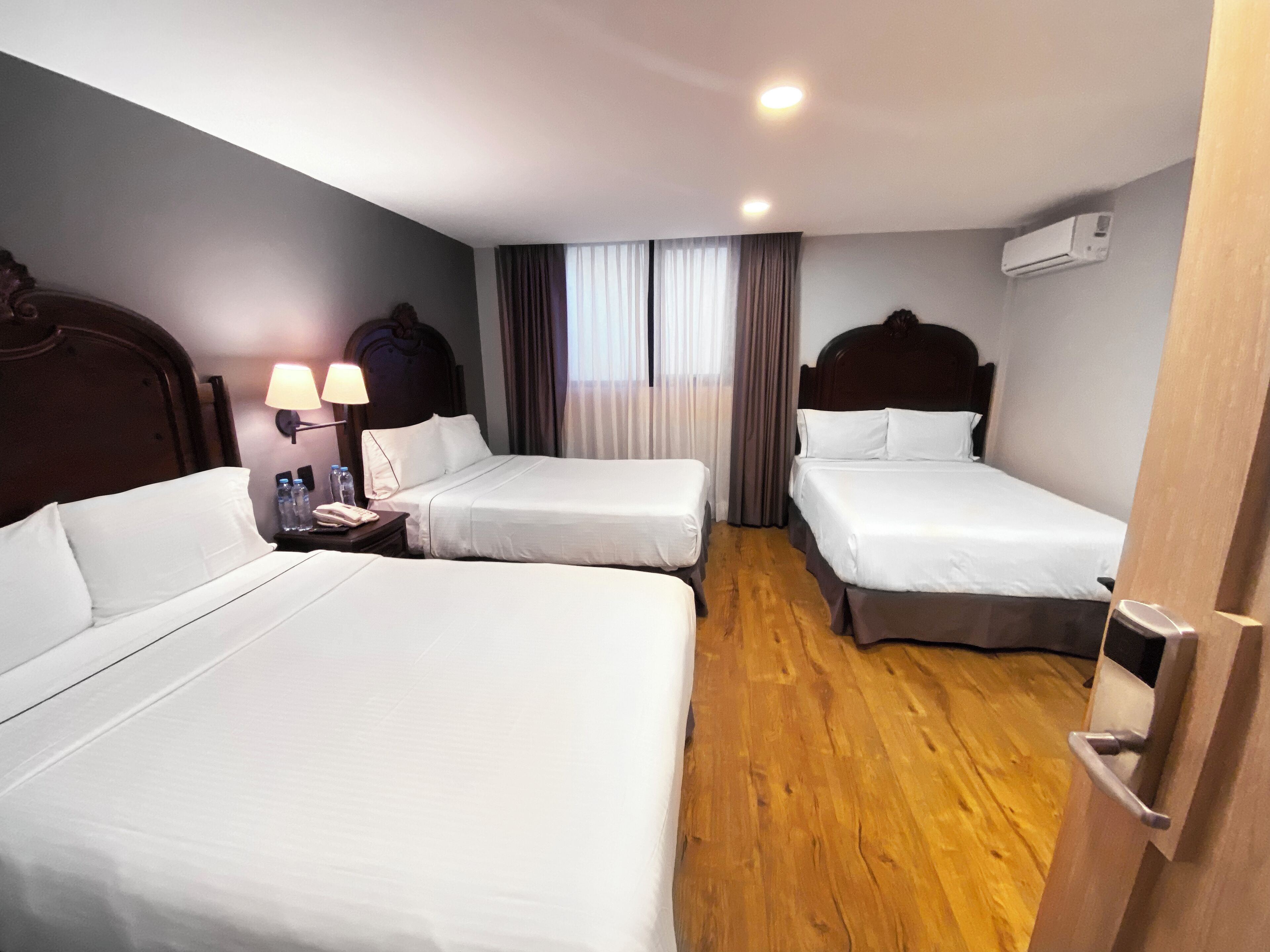 Una habitación de hotel con dos camas individuales, suelo de madera, una mesita de noche con lámpara y un equipo de aire acondicionado en la pared.