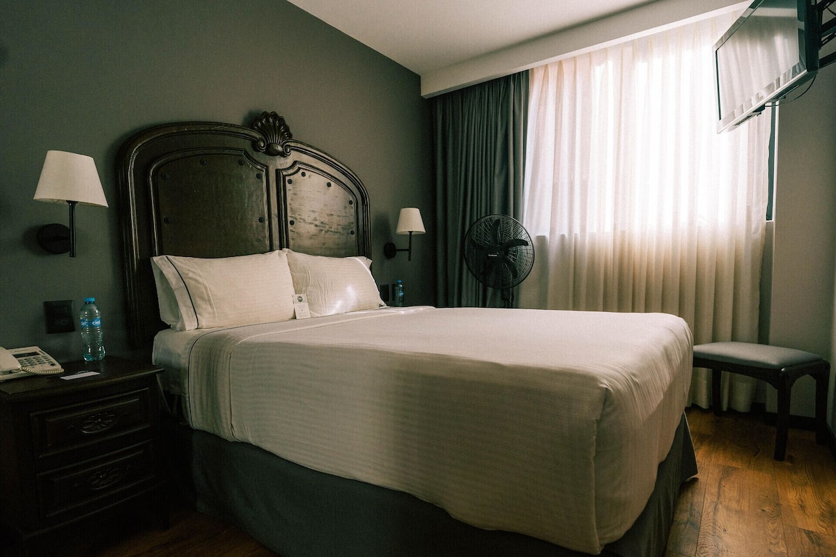 A neatly made bed with a headboard, a bedside table with a lamp, a water bottle, and a chair.