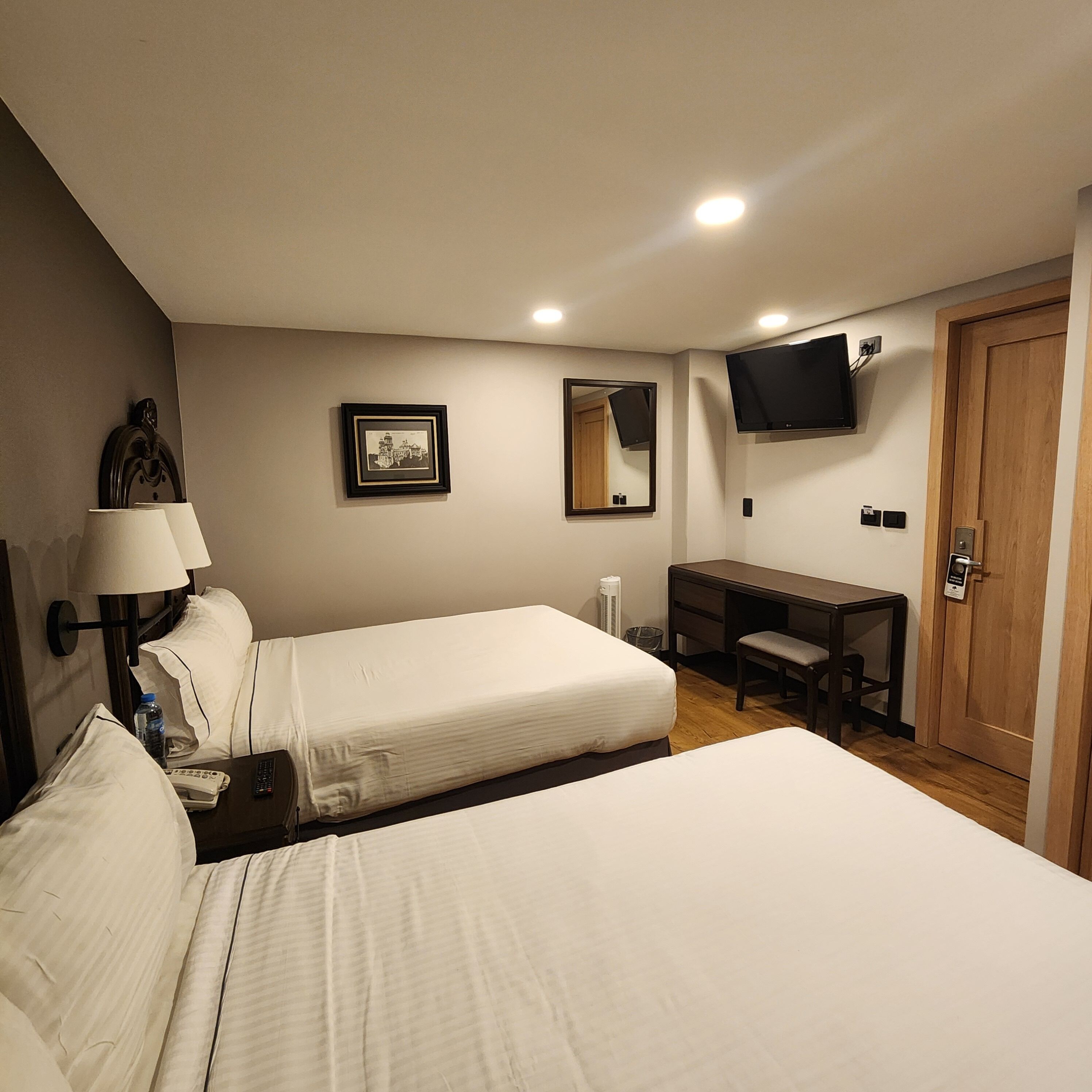 A hotel room with two beds, a desk, a chair, a TV, a mirror, and a door.