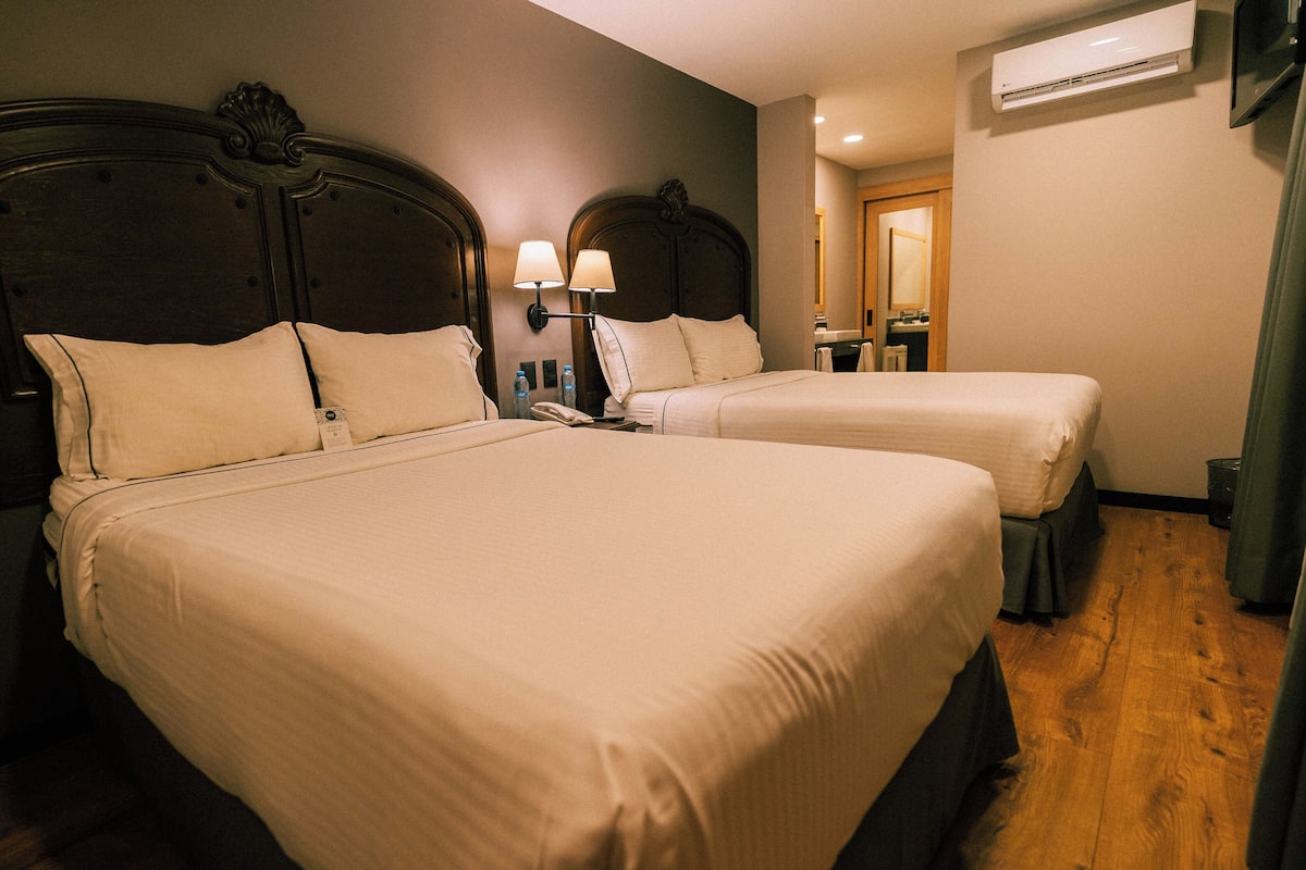 A hotel room with two beds, a nightstand with a lamp, an air conditioning unit, and a wooden floor.