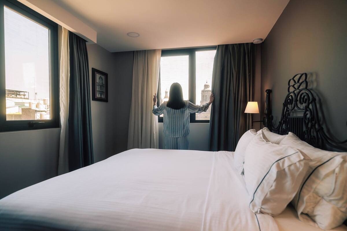 A hotel room with a large bed, a window with curtains, and a view of a cityscape.