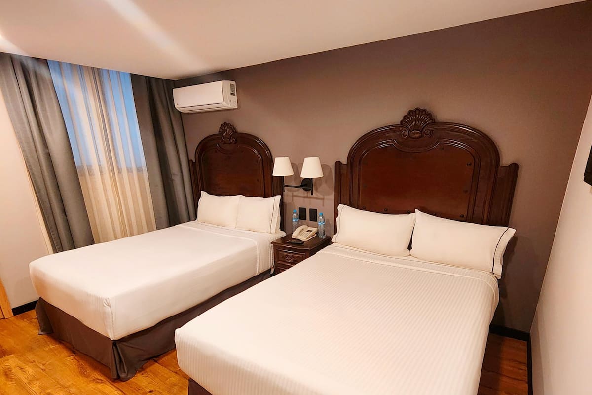 A hotel room with two beds, wooden flooring, a nightstand with a lamp, and an air conditioning unit on the wall.