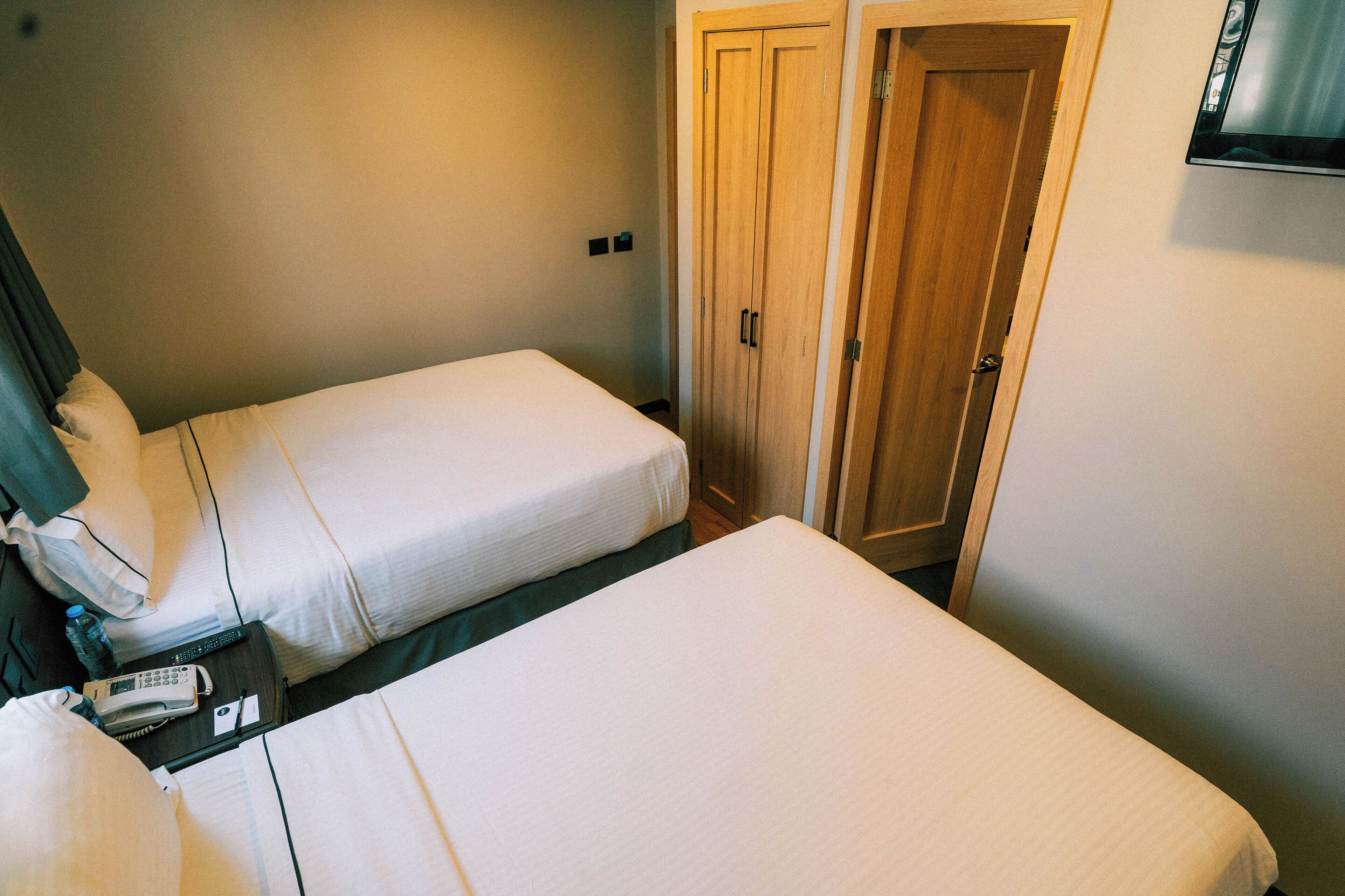 Une chambre d’hôtel avec deux lits, une table de chevet avec un téléphone et une bouteille, et une armoire en bois.