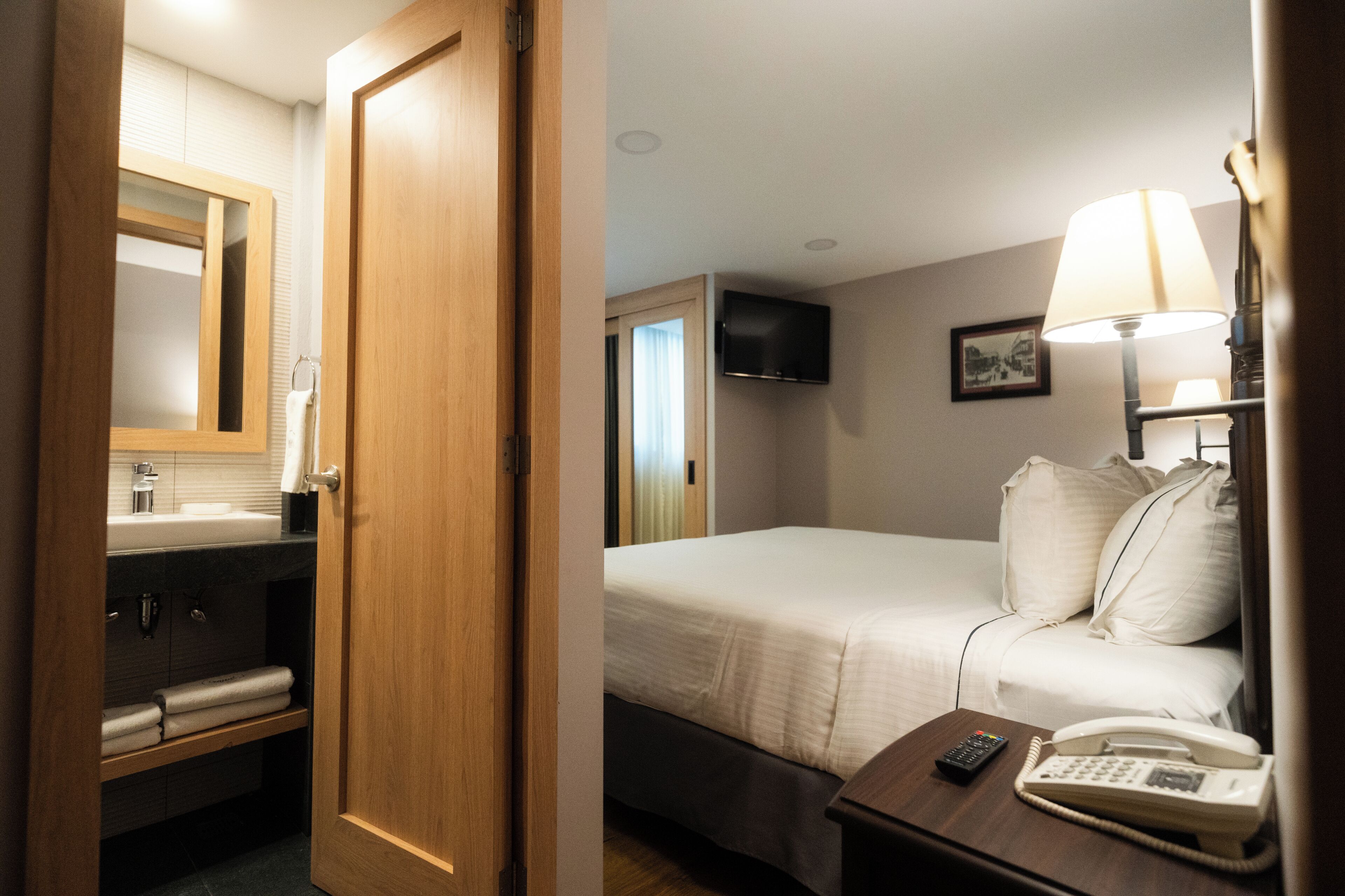 A hotel room with a wooden door, a bed with white bedding, a bedside table with a lamp and a telephone, a TV mounted on the wall, and a bathroom with a mirror, sink, and towel rack.