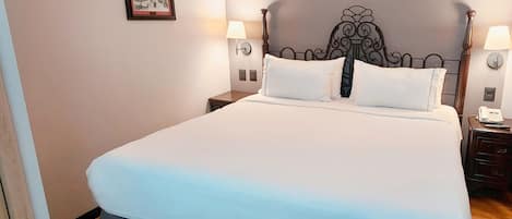 A neatly made bed with a white and blue bedspread, two bedside tables with lamps, a framed picture on the wall, and a wooden floor.