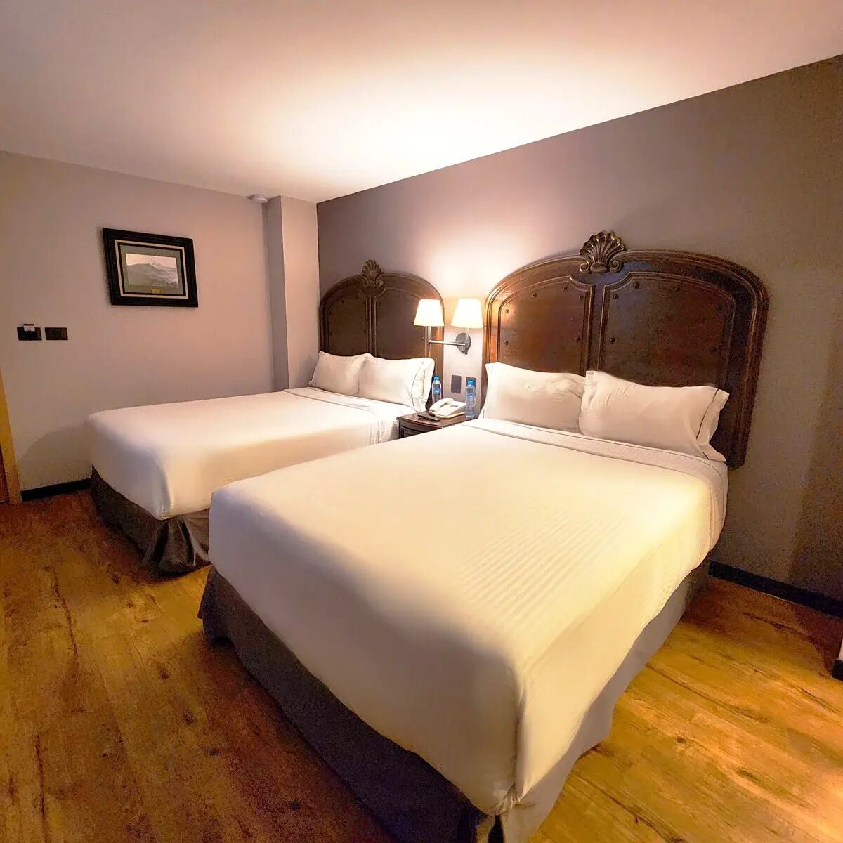 Habitación de hotel con dos camas individuales, cabecera de madera, mesita de noche con lámpara y un cuadro en la pared.