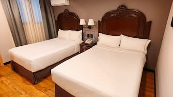 A hotel room with two beds, wooden flooring, a nightstand with a lamp, and an air conditioning unit on the wall.