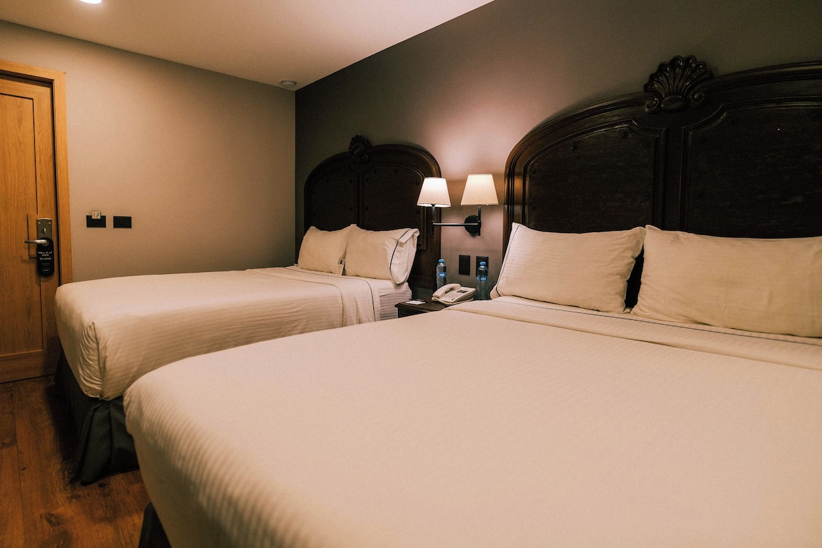 A hotel room with two beds, a wooden door, a nightstand with a lamp, and a wall-mounted phone.