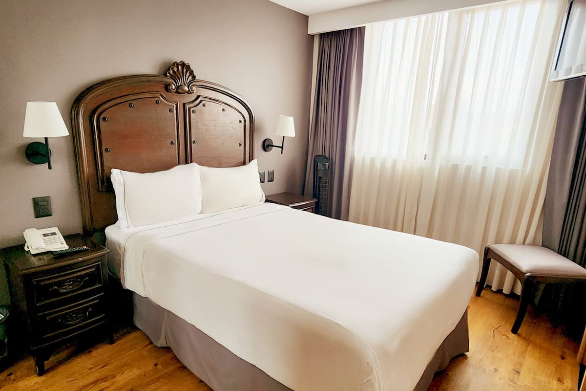A hotel room with a large bed, wooden headboard, two bedside tables with lamps, a small chair, and a window with curtains.