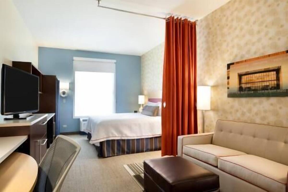 A hotel room with a bed, a desk with a computer, a sofa, a TV, and a window with curtains.