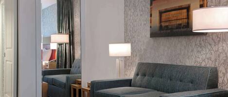 A hotel room with a grey sofa, a small table, a lamp, and a mirror reflecting the room.