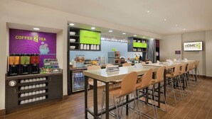 A hotel breakfast area with a coffee station, a refrigerator with beverages, and a long table with chairs.